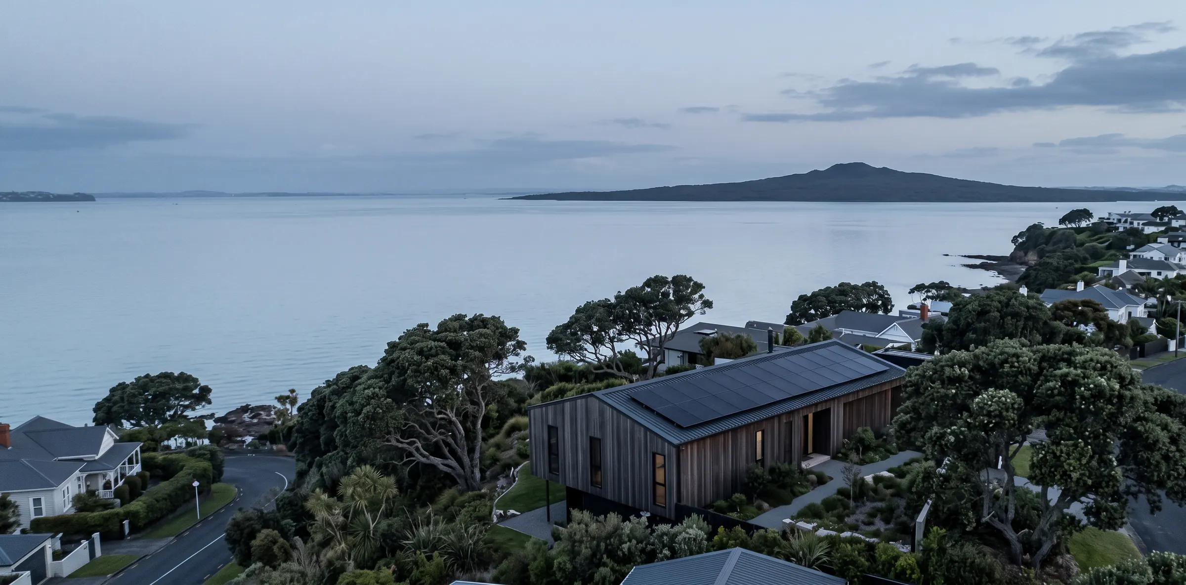Aerial view at dawn over Devonport, a dark-timber home with a rooftop solar array, the Waitematā Harbour and Rangitoto beyond
